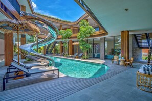 A modern indoor pool area with a spiral slide, lounge chairs, and a wooden deck. - Akashi Residence (Canggu)
