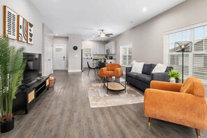 Una sala moderna con un sofá, sillones y una mesa de centro. Se observa una cocina al fondo con armarios blancos y un comedor con sillas.