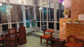 A cozy indoor seating area with wooden chairs and tables, large windows with a view of the outside, and a brick wall with a small shelf holding plants. - Mary Land  Homestay - A  "Private Pool Villa" Near Trivandrum Airport and Beach. (Thiruvananthapuram)