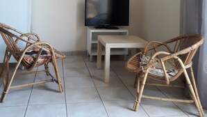 A small living area with two wicker chairs, a television on a white stand, a small table, and a refrigerator.