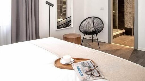 A modern hotel room with a large bed, a reading chair, a small table with a cup and glasses, and a door leading to a bathroom. - N-SPACE MODERN GUEST ROOMS (Rome)