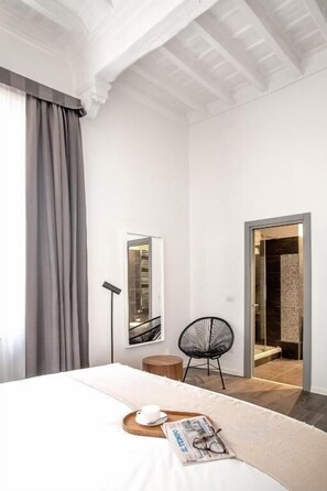 A modern hotel room with a large bed, a reading chair, a small table with a cup and glasses, and a door leading to a bathroom.