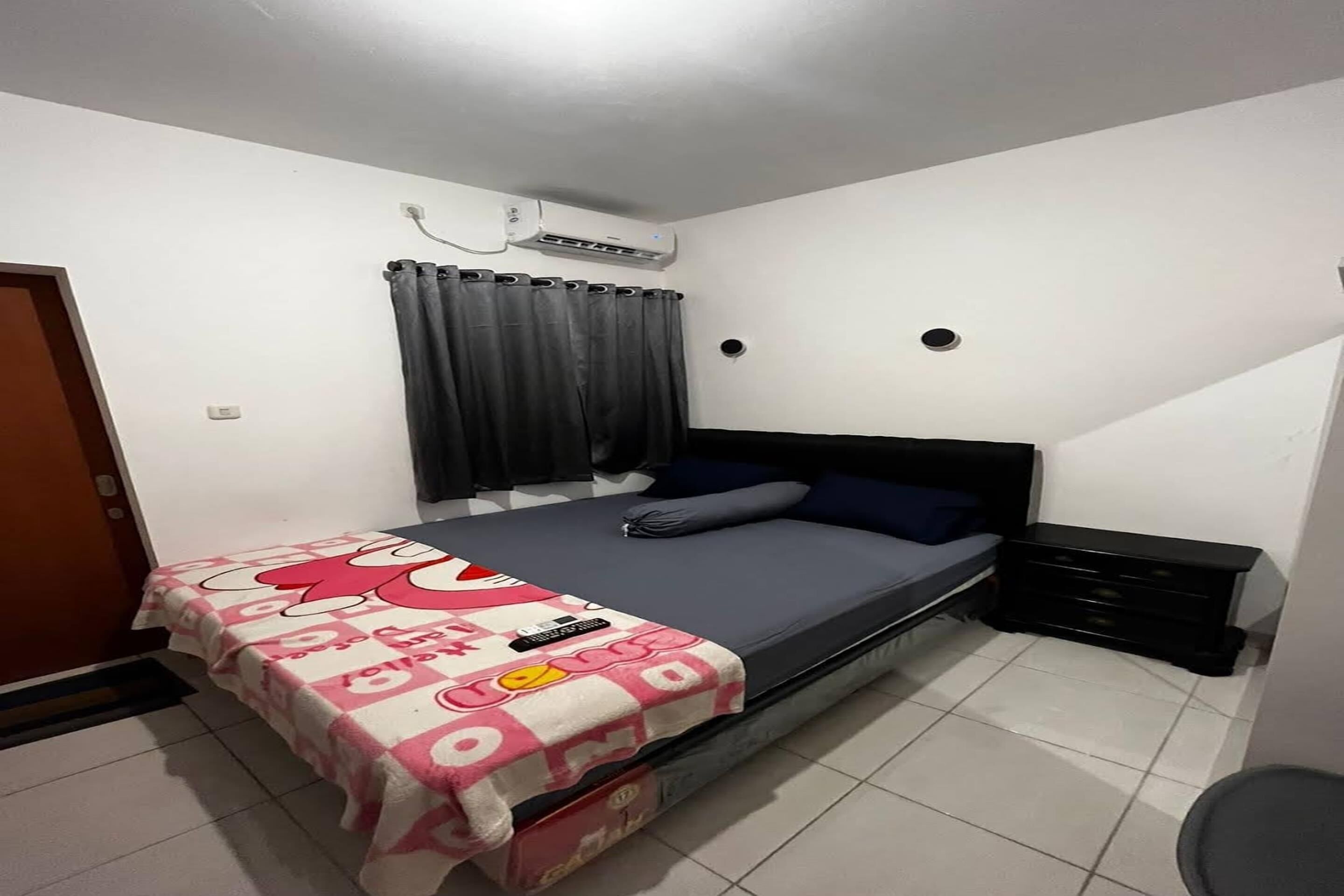 Een slaapkamer met een bed, nachtkastje, airconditioning, gordijn en een deur.