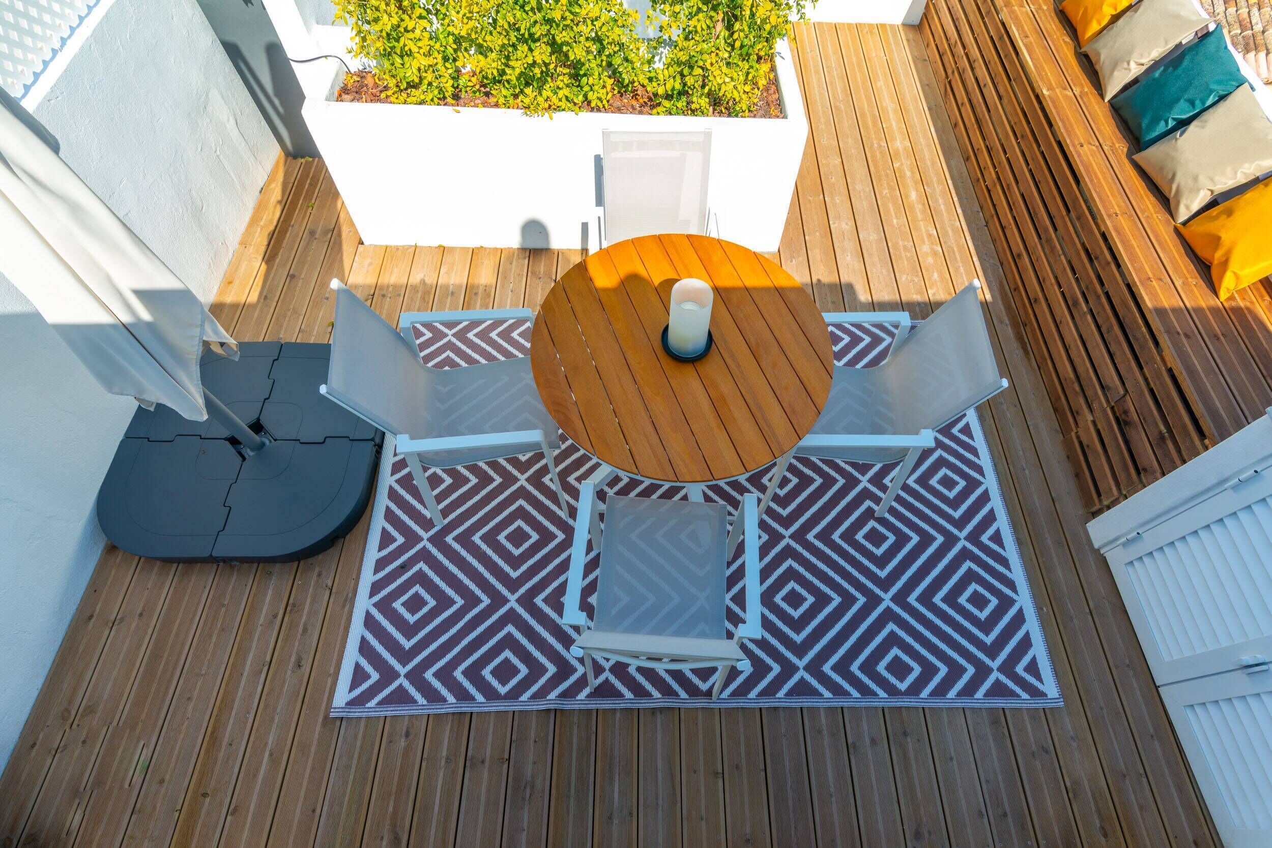 A wooden deck with a round table and two chairs, a patterned rug, and a white planter with green foliage.