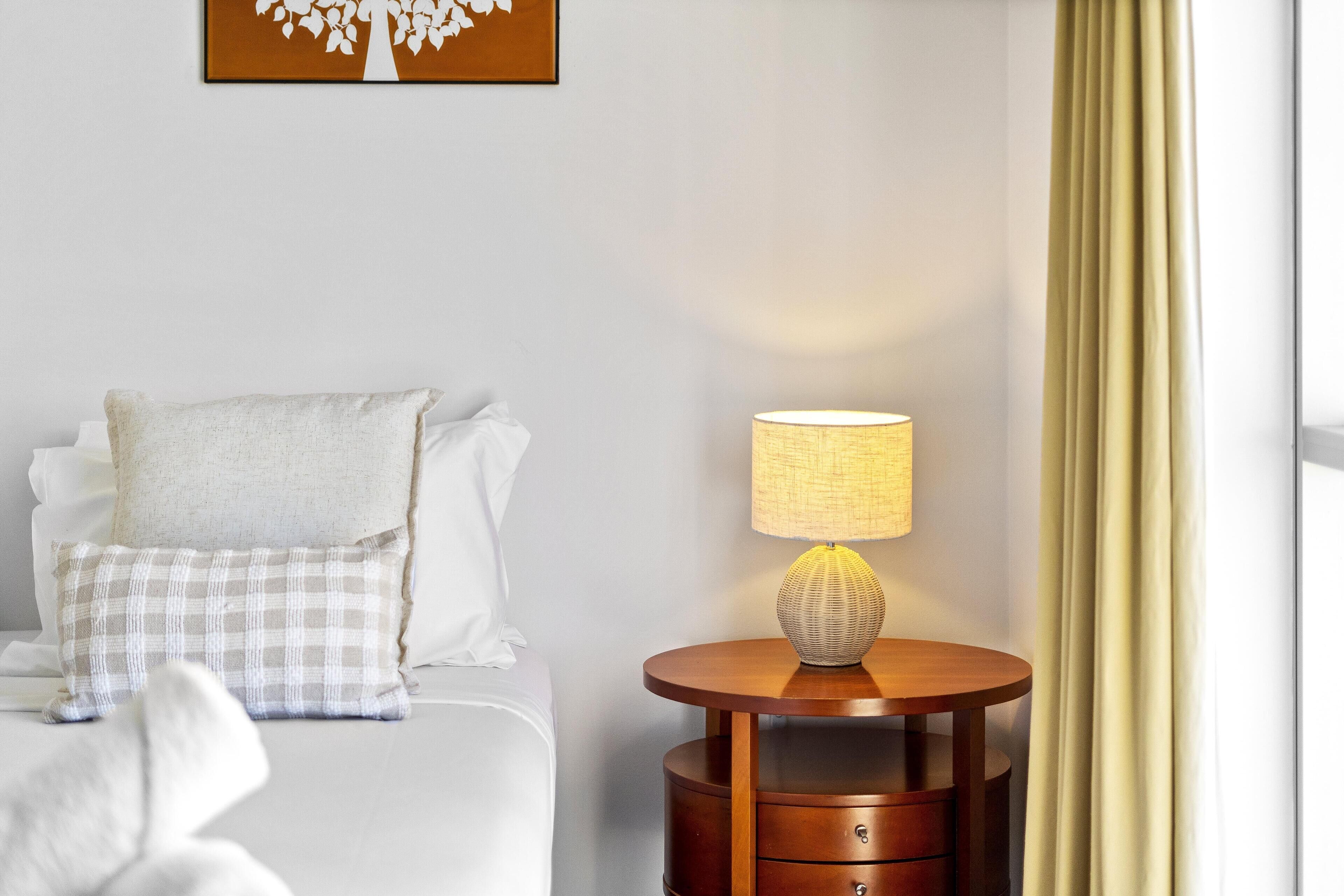 A neatly made bed with a white and plaid pillow, a round wooden nightstand with a lamp, and a framed picture on the wall.