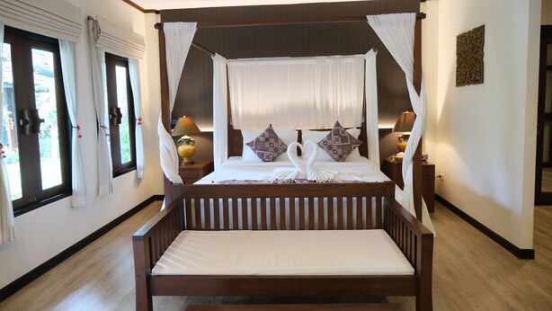 A four-poster bed with a canopy, two bedside tables with lamps, and a view of the outdoors through large windows. - Vimarn Kaew Village and Resort (Chiang Mai)