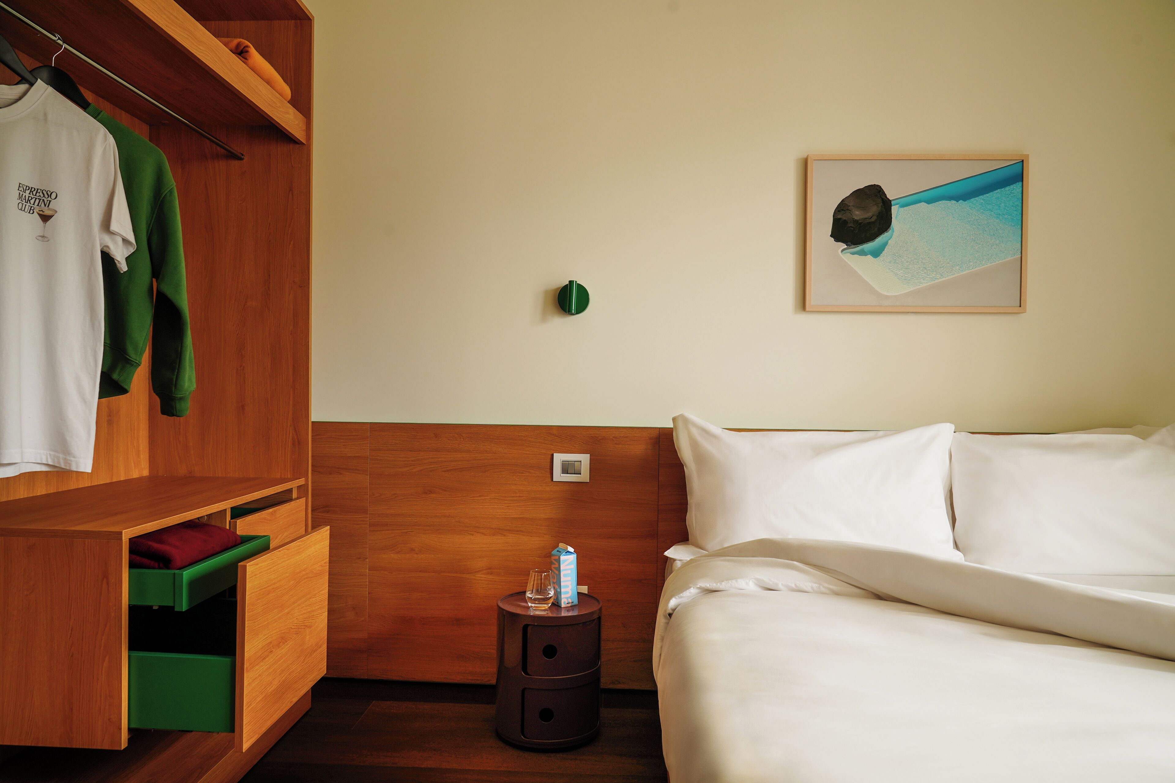 A hotel room with a bed, a wooden wardrobe, a small bedside table with a glass and a can, and a painting on the wall.