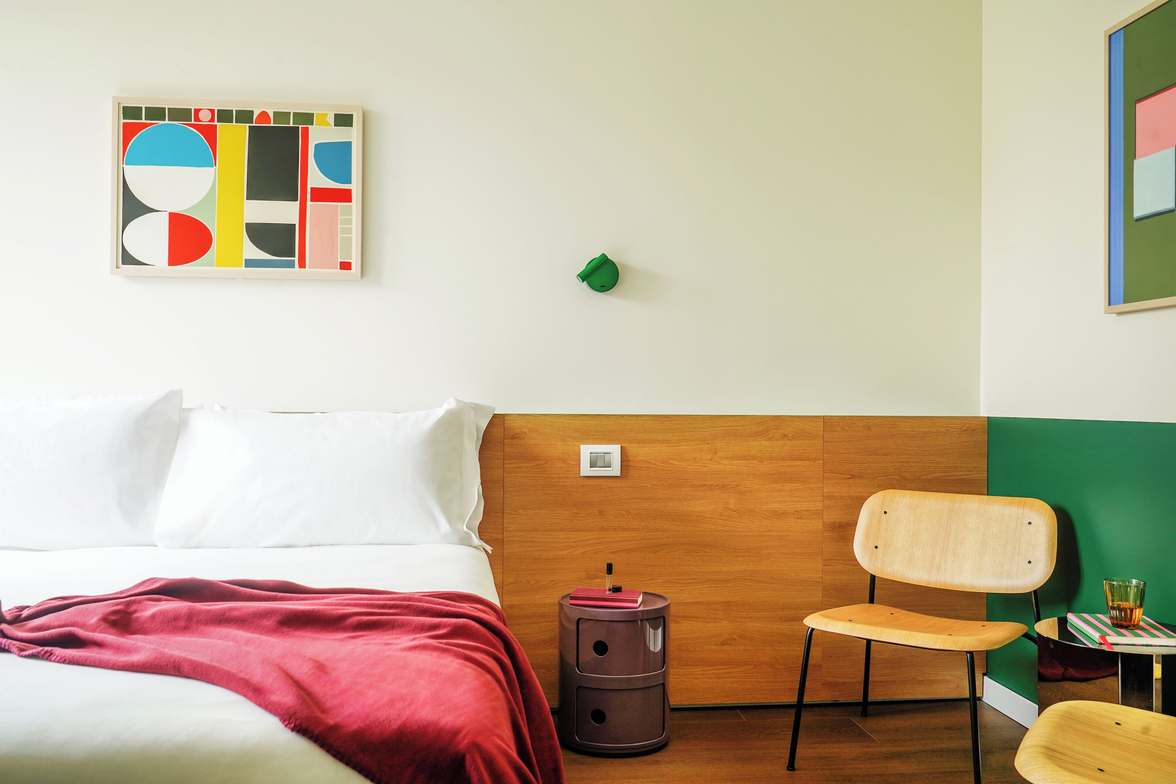 Ein modernes Hotelzimmer mit einem hölzernen Kopfteil, einem Bett mit weißen Leinen, einer roten Tagesdecke, einem kleinen runden Tisch mit einem Glas und einem Stuhl.