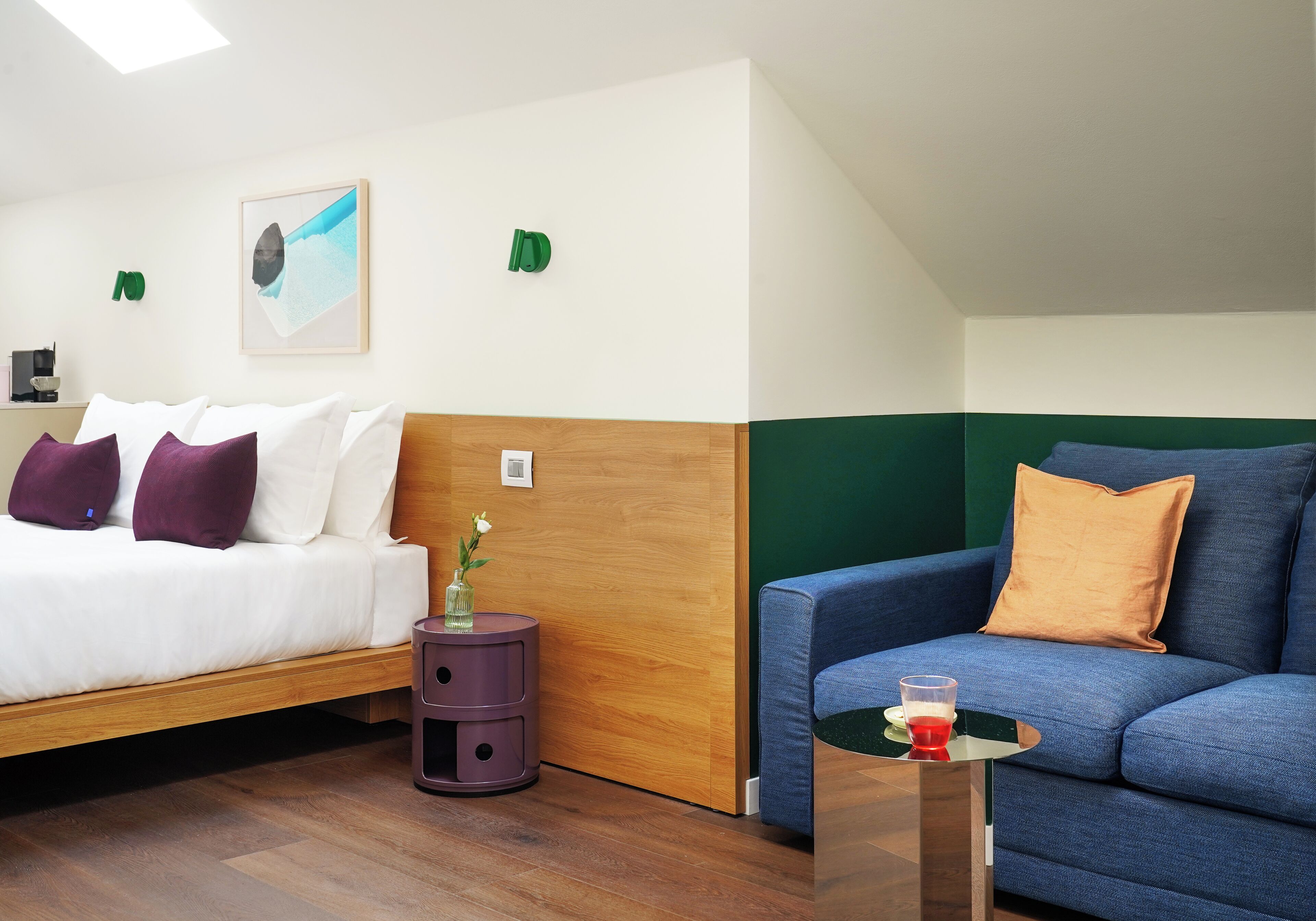 Une chambre d’hôtel avec un lit, un canapé bleu, une petite table avec un verre et une tête de lit en bois.