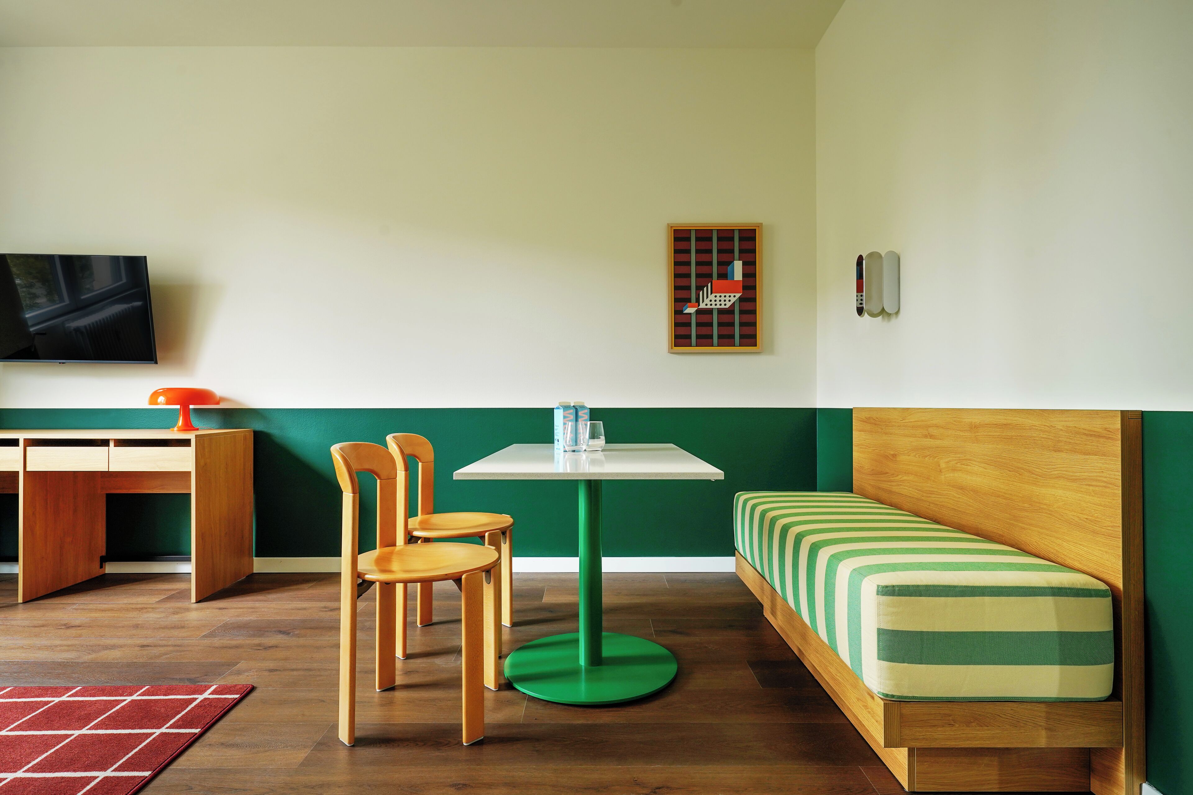 Une pièce avec un bureau en bois, deux chaises, une petite table avec une lampe, un lit avec une literie à rayures et un téléviseur fixé au mur.