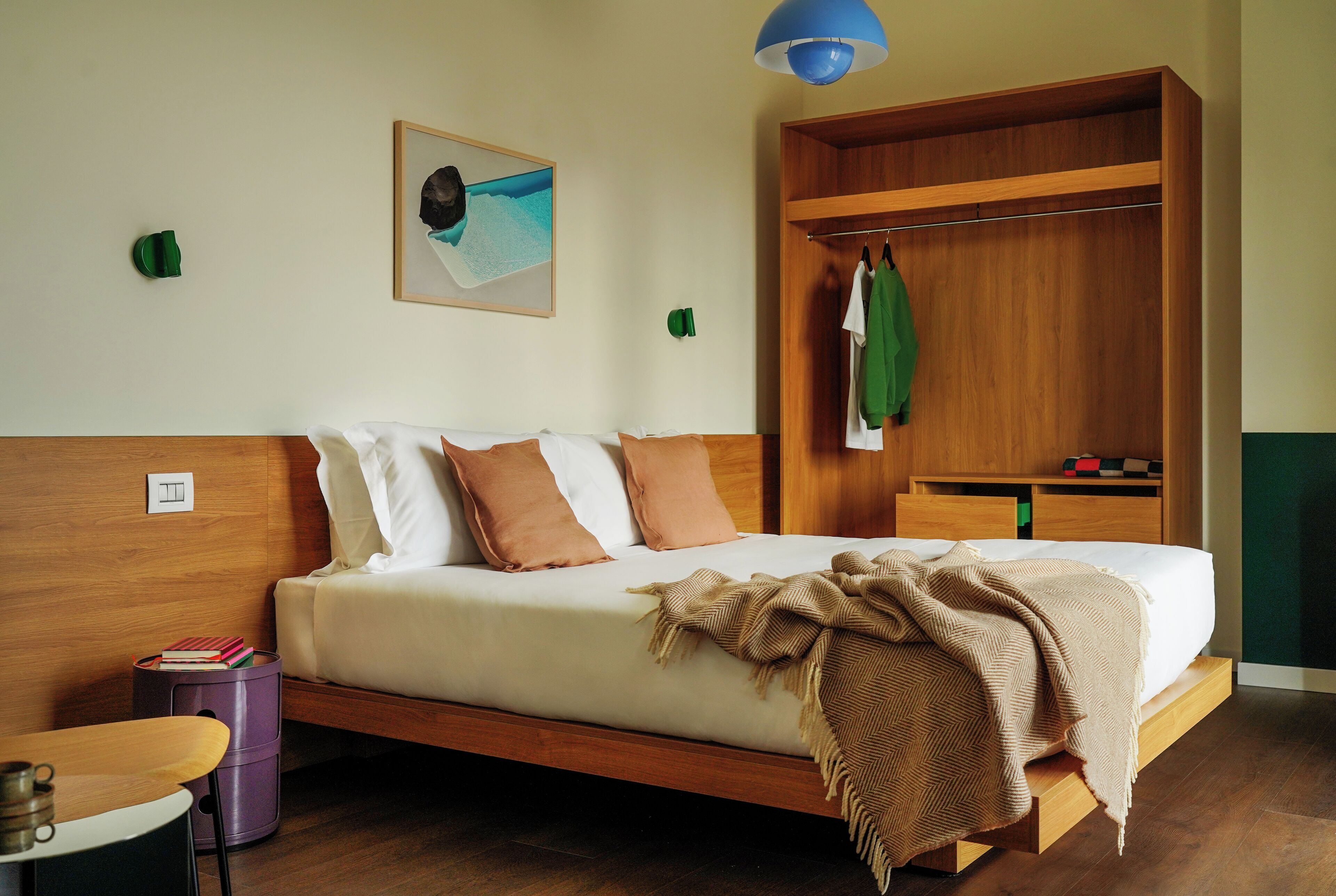 A modern bedroom with a wooden bed, a small desk, a chair, a wardrobe, and a painting on the wall.