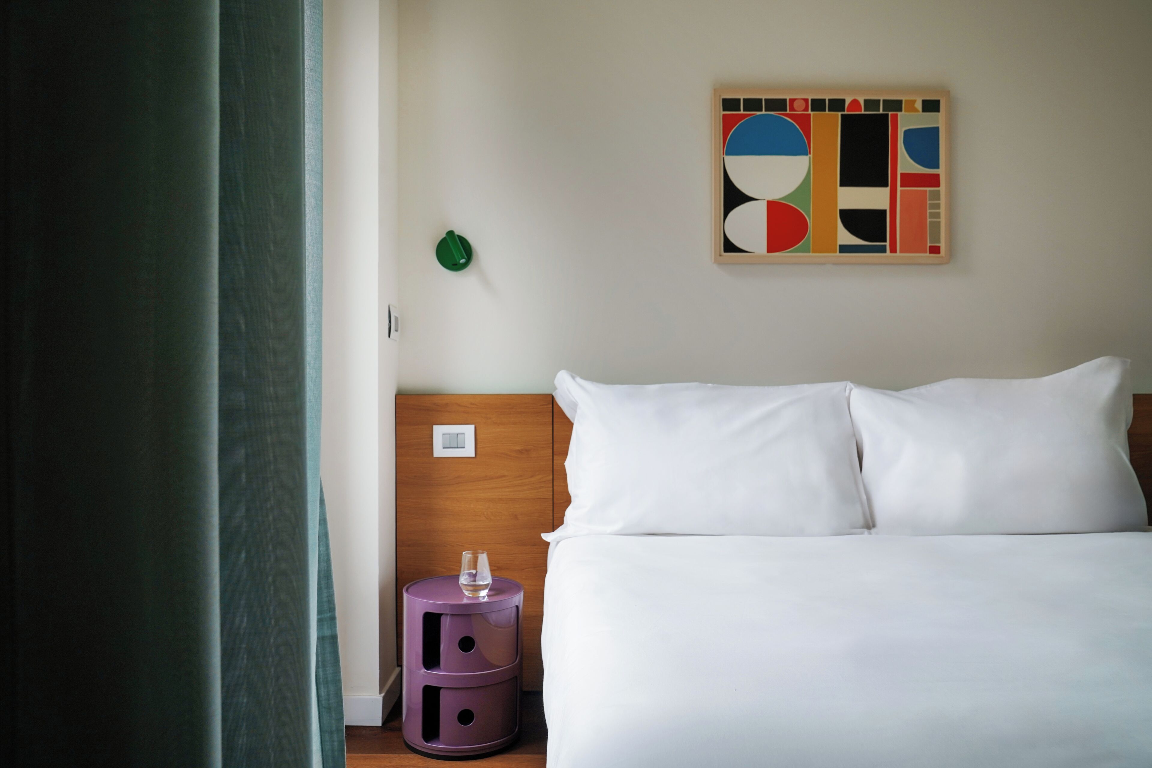 A hotel room with a bed, a nightstand, a wall art piece, and a green curtain.
