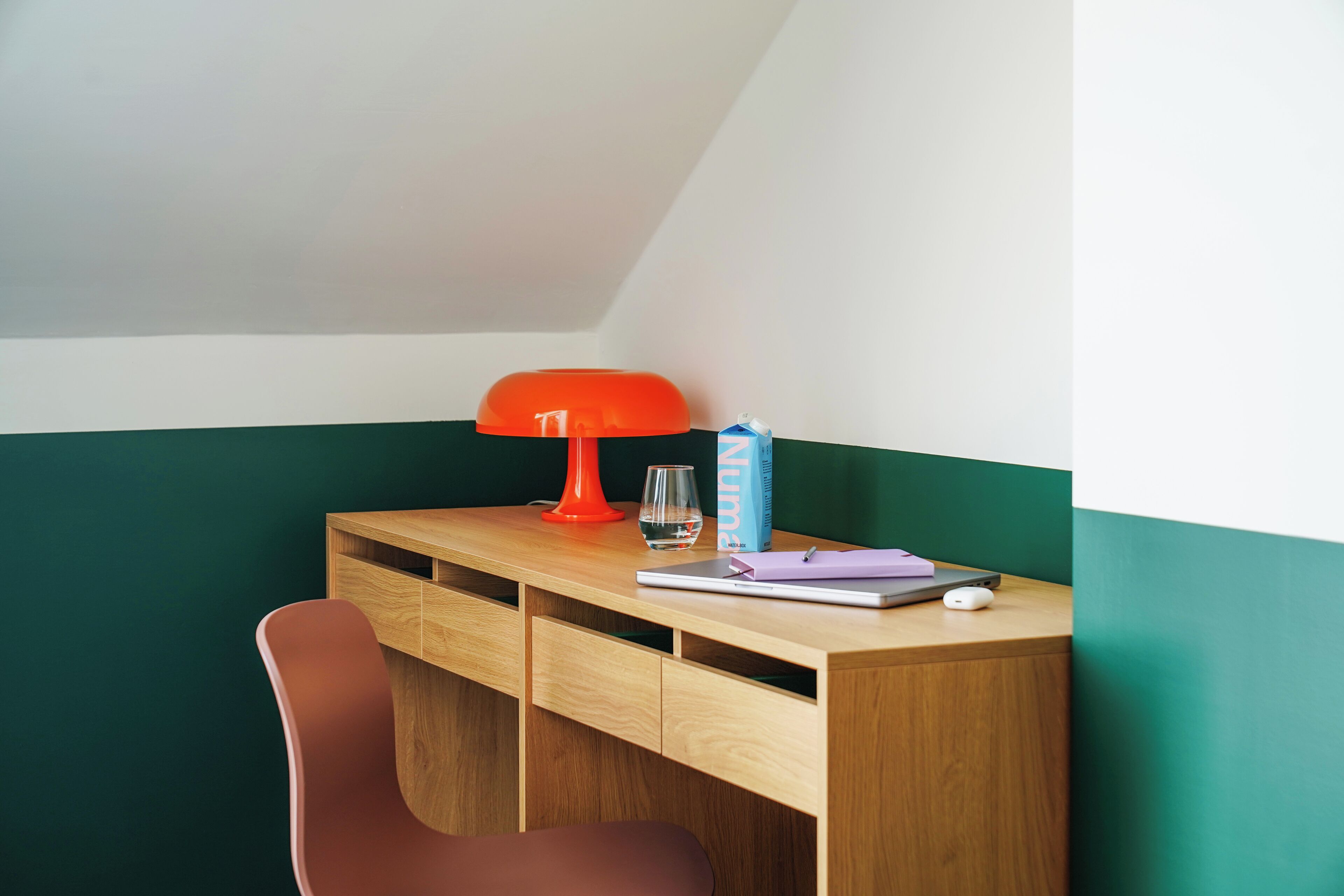 A study area with a wooden desk, a red lamp, a glass of water, a notebook, and a pen.