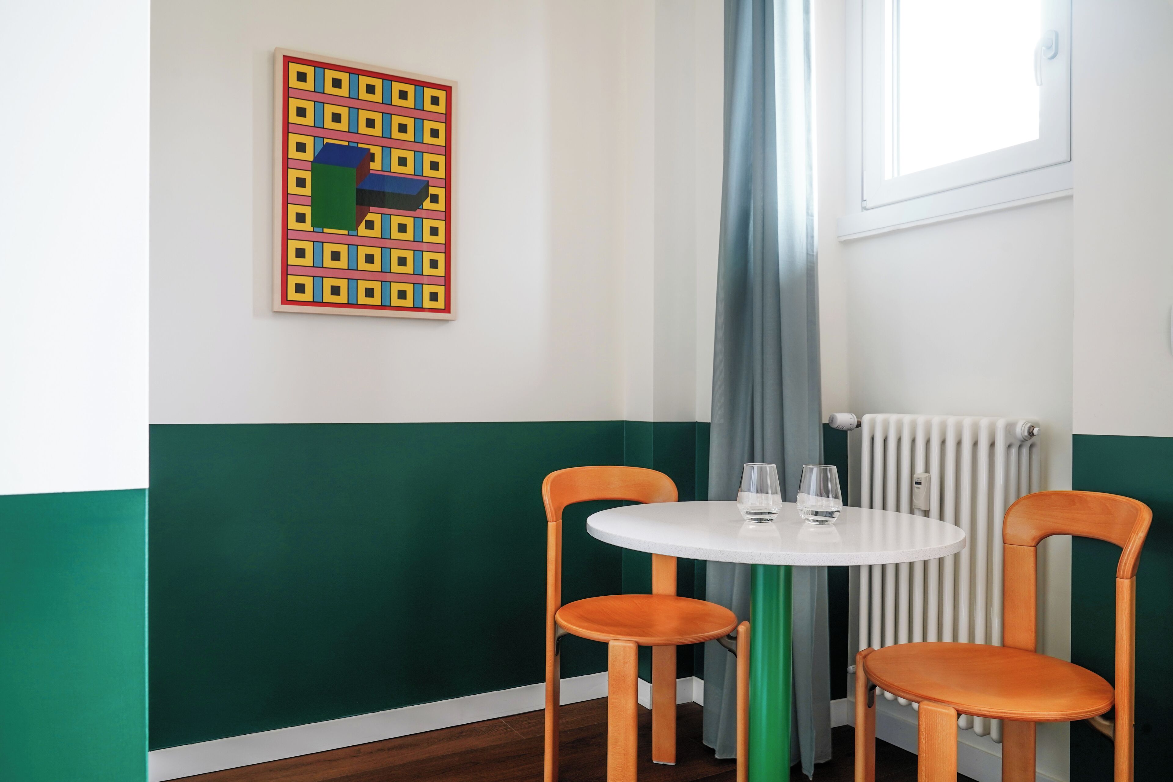 A room with a round white table, two wooden chairs, a radiator, a window with curtains, and a colorful abstract painting on the wall.