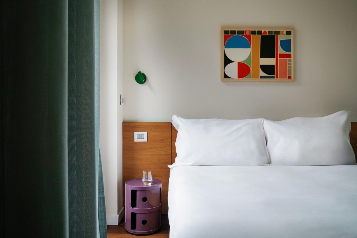 A hotel room with a bed, a nightstand, a wall art piece, and a green object on the wall.