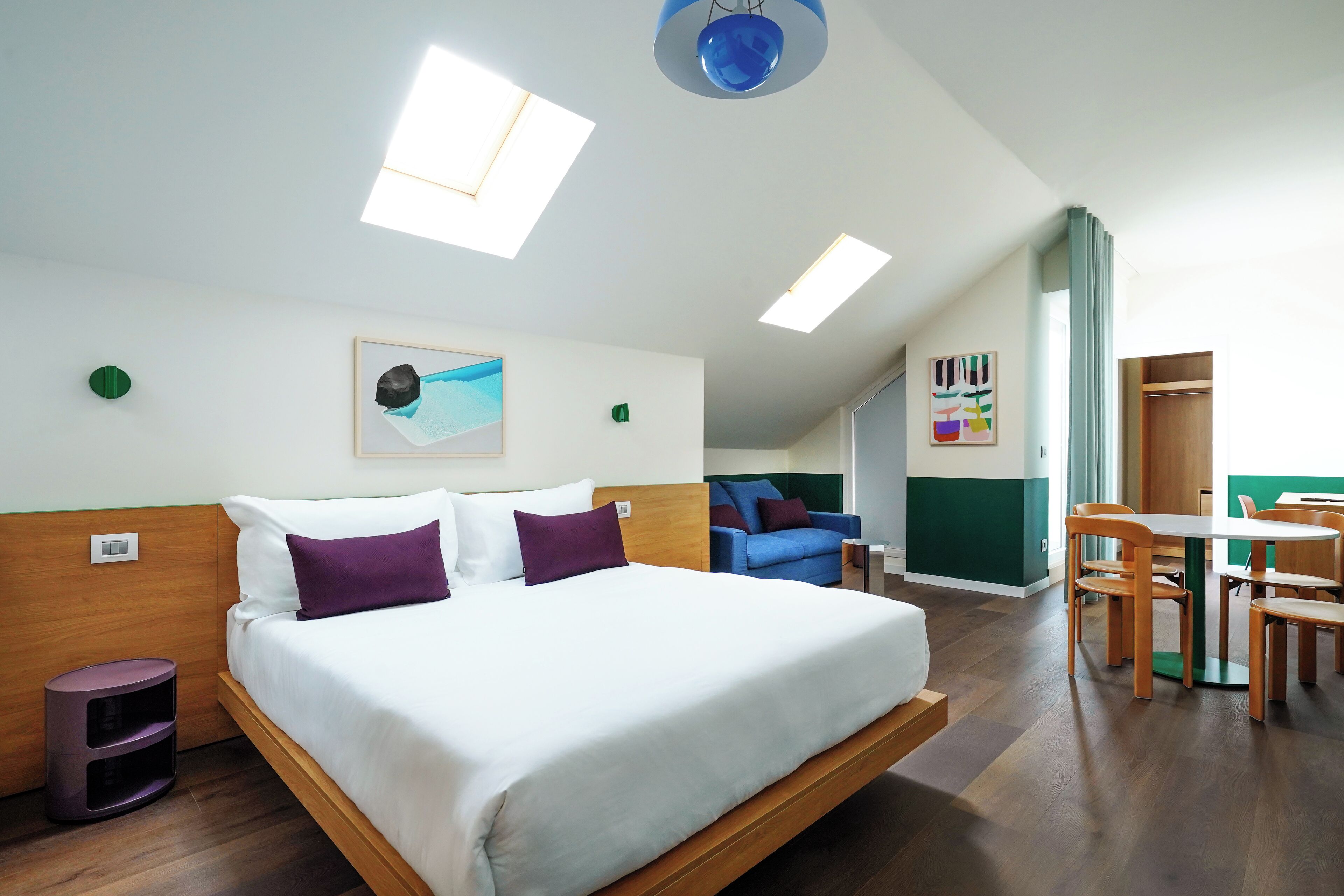 A modern hotel room with a large bed, a dining table with chairs, and a blue ceiling light.