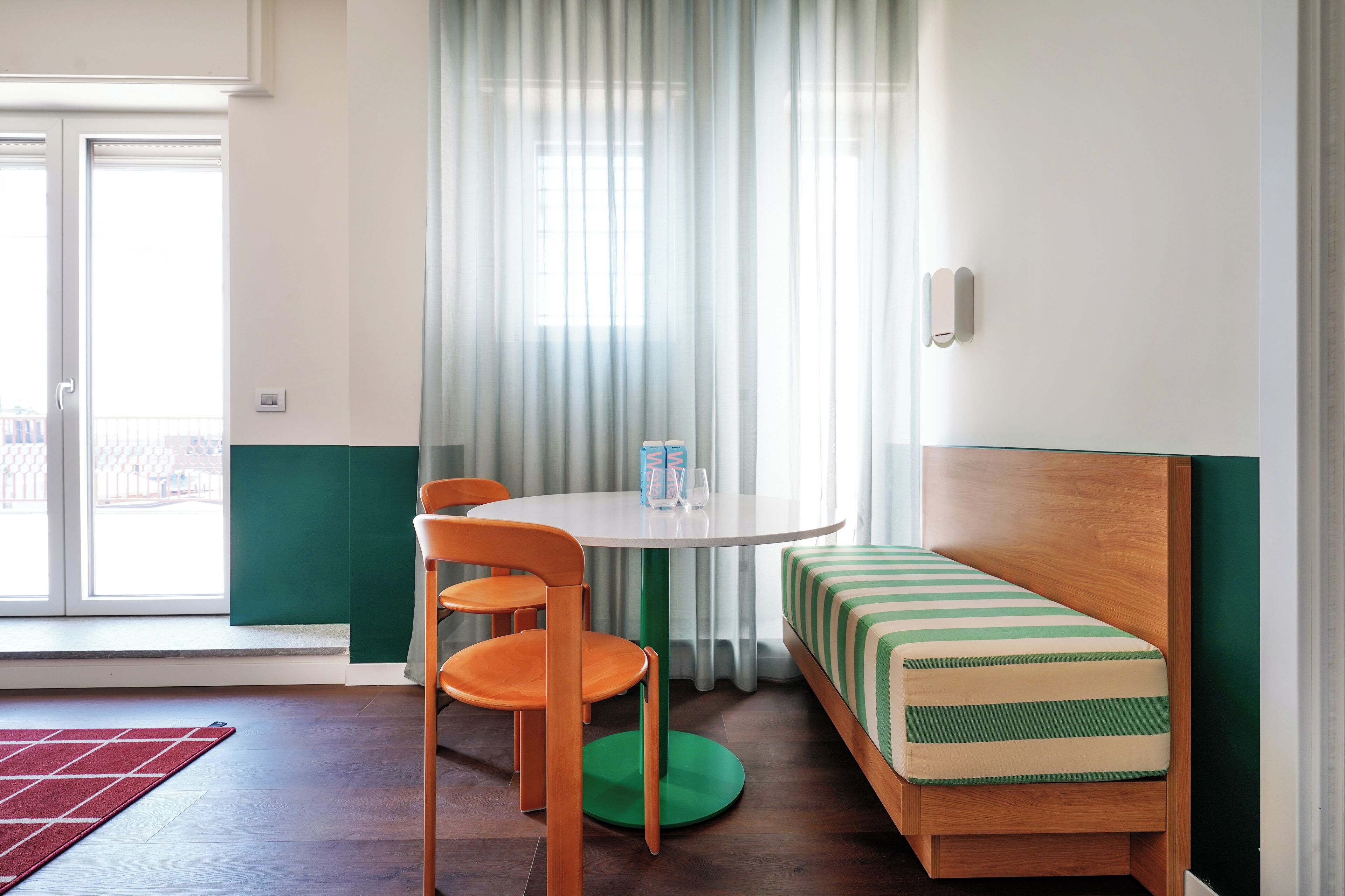 Une pièce avec une table ronde, deux chaises orange, un tapis vert, un banc à rayures et une fenêtre ornée de rideaux légers.