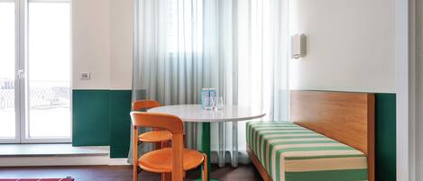 Une pièce avec une table ronde, deux chaises orange, un tapis vert, un banc à rayures et une fenêtre ornée de rideaux légers.