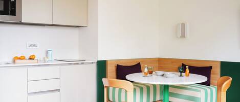 A compact kitchen with a microwave, a sink, and a countertop with a few items. There is a small dining area with a round table and two chairs, and a bench with a striped cushion.
