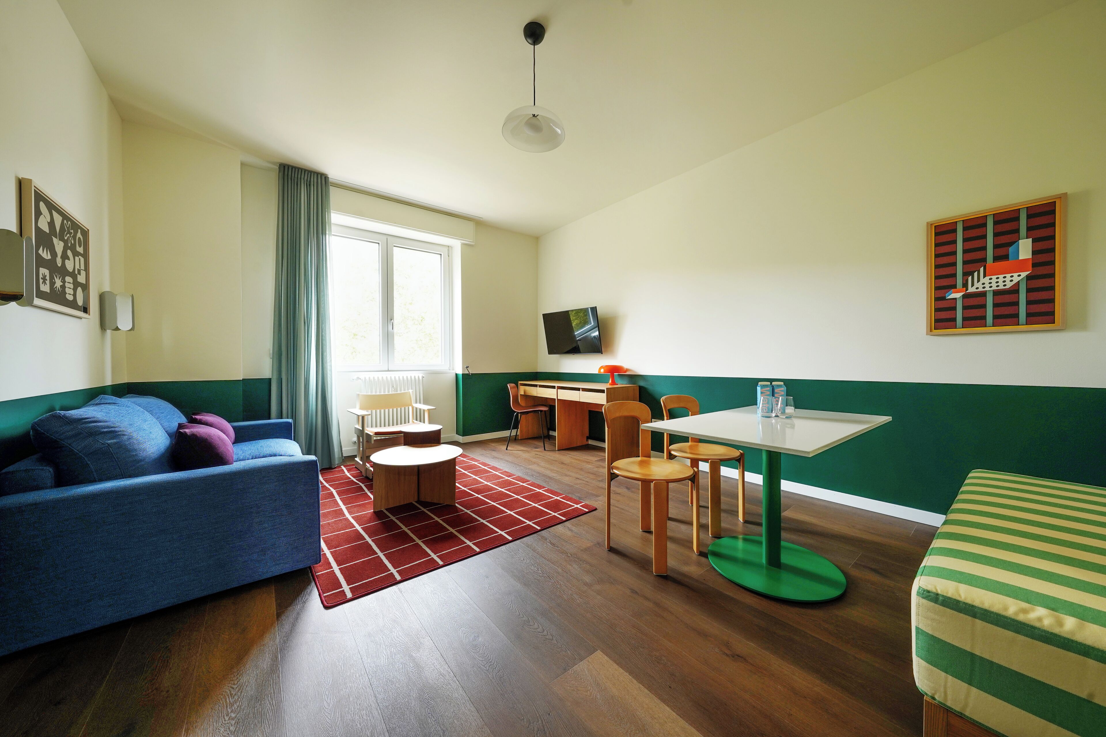 A room with a blue sofa, a green-striped bed, a dining table with chairs, a TV on the wall, and a window with curtains.