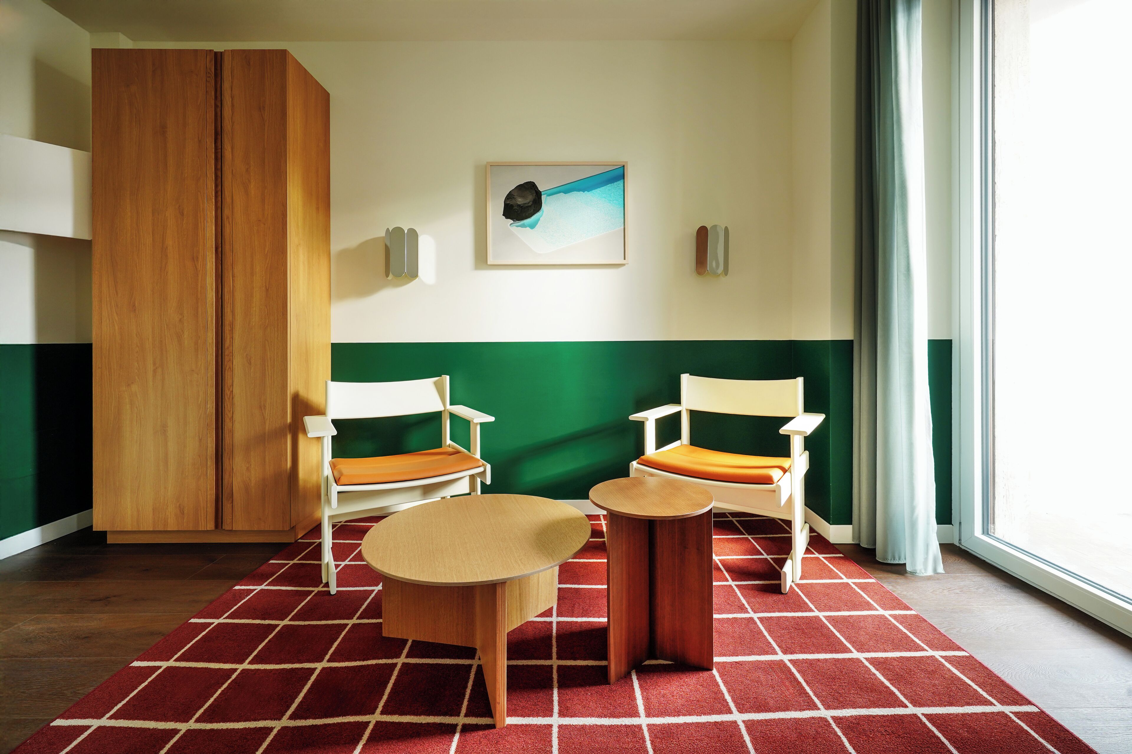 A hotel room with a wooden wardrobe, two white chairs with orange cushions, a small round table, a red and white checkered rug, and a painting on the wall.