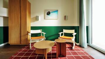 Una camera d'albergo con un'armadio in legno, due sedie bianche con cuscini arancioni, un piccolo tavolino rotondo, un tappeto a scacchi rosso e bianco e un quadro appeso al muro.