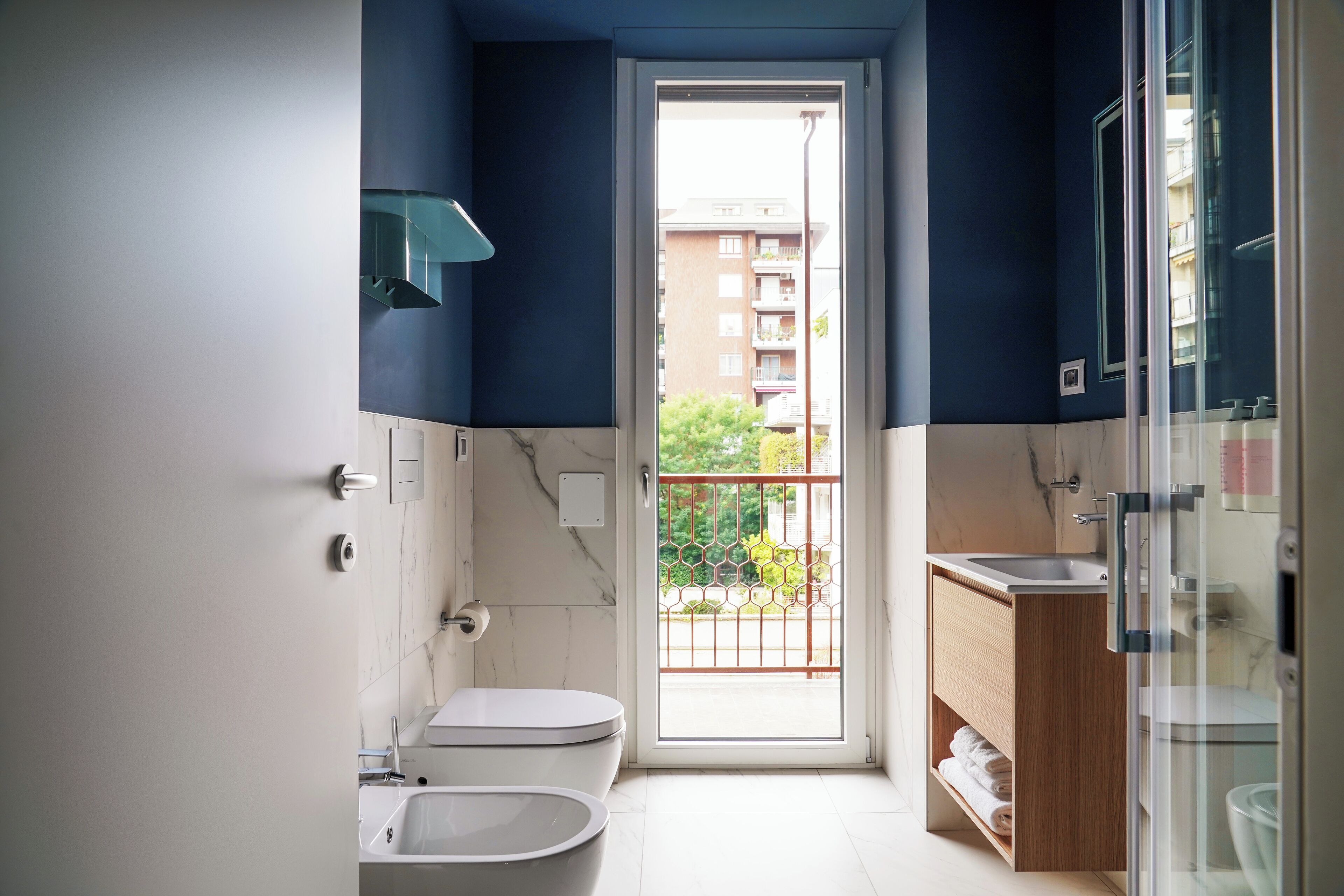 A compact bathroom with a toilet, bidet, and sink, featuring a view of a balcony and a building outside.