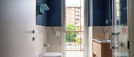Et lille badeværelse med toilet, bidet og håndvask, med udsigt til en balkon og en bygning.