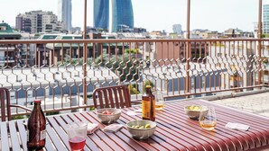 Une table avec une bouteille, des verres, des bols et un menu, sur un balcon donnant sur un paysage urbain avec des bâtiments modernes.
