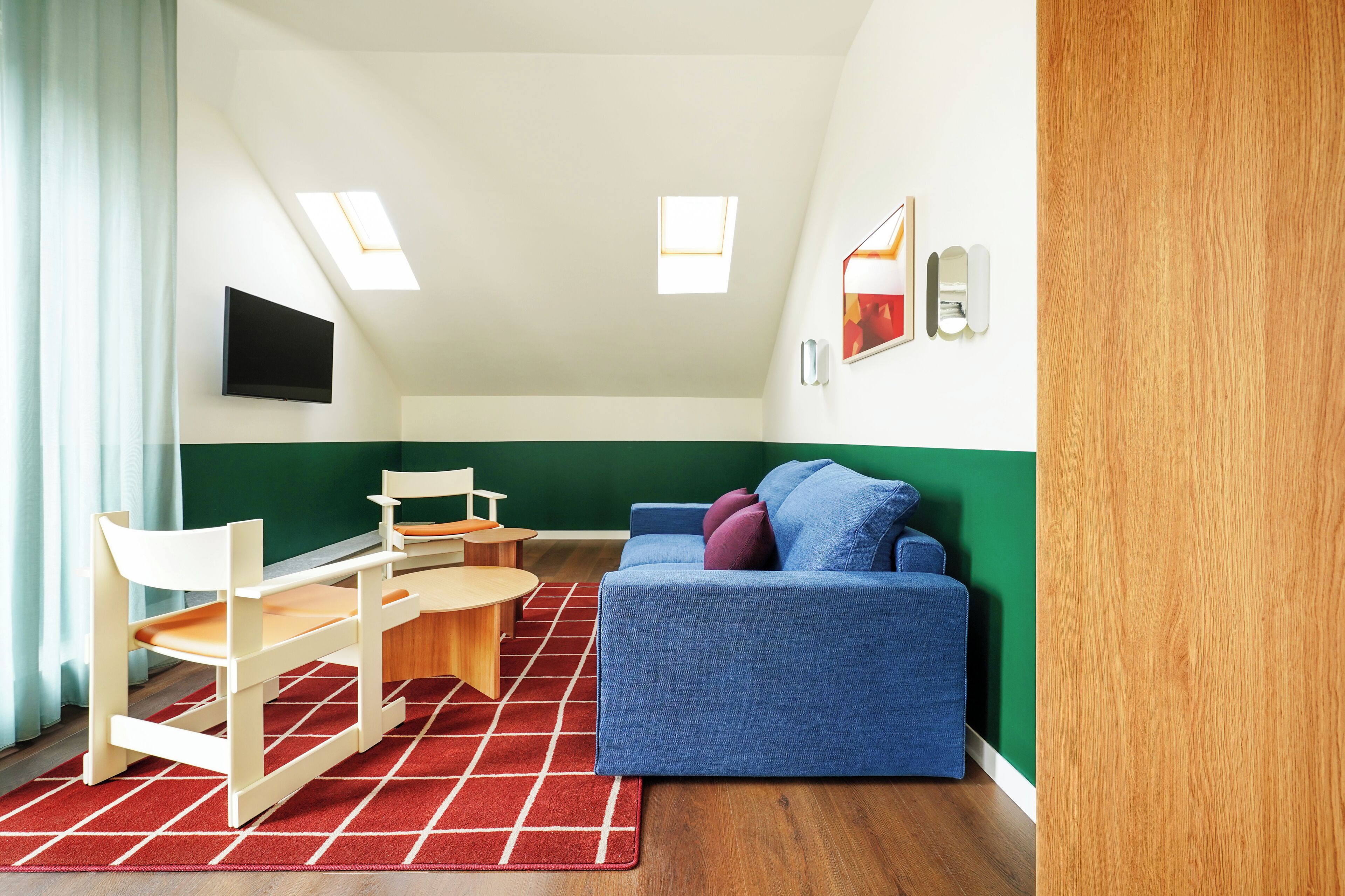 A room with a blue sofa, wooden chairs, a small table, and a red rug. There are skylights on the ceiling and a green accent wall.