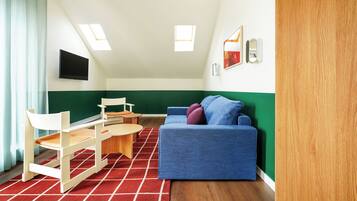 A room with a blue sofa, wooden chairs, a small table, and a red rug. There are skylights on the ceiling and a green accent wall.