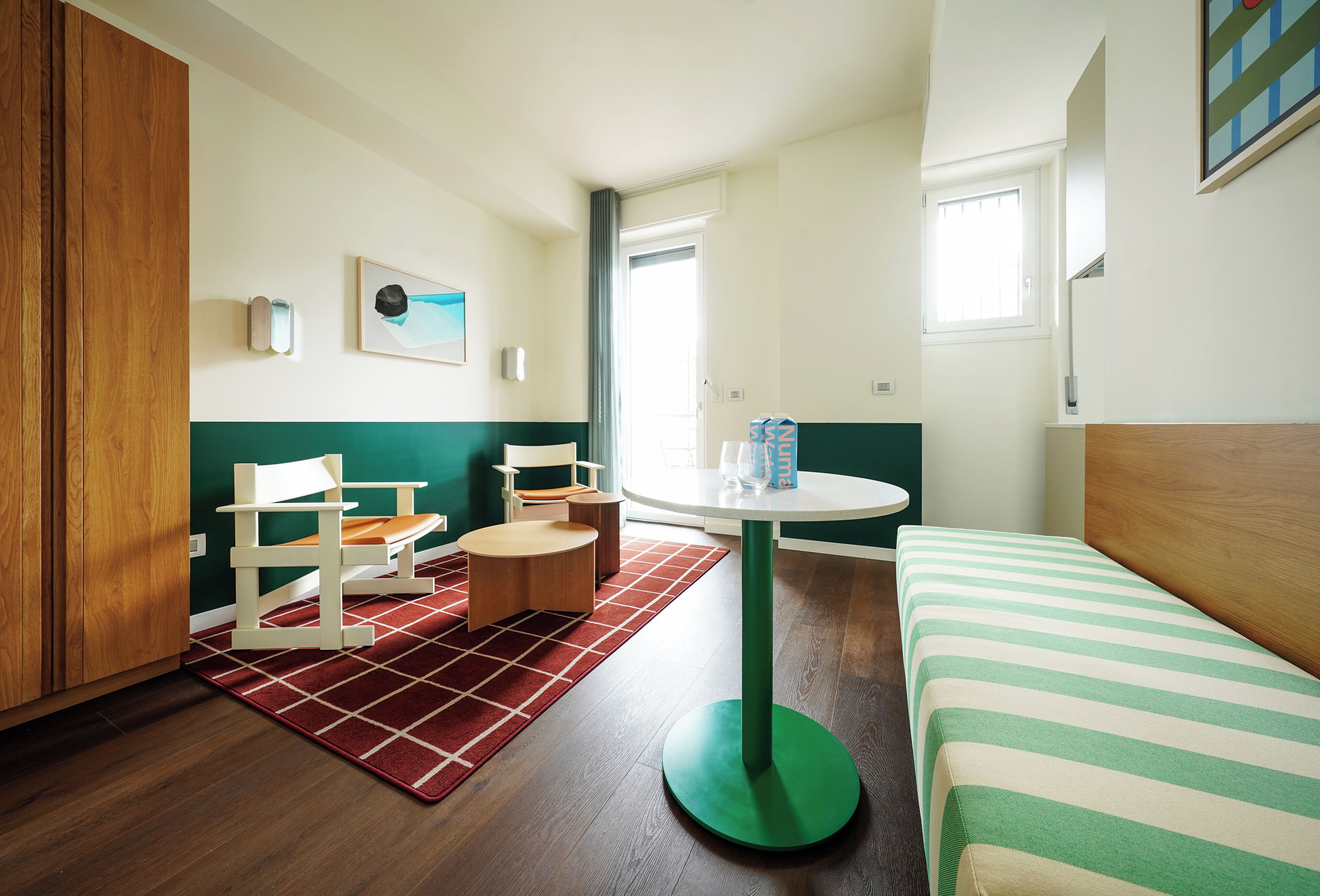 A hotel room with a bed, a round table, a chair, a small table, and a red rug.