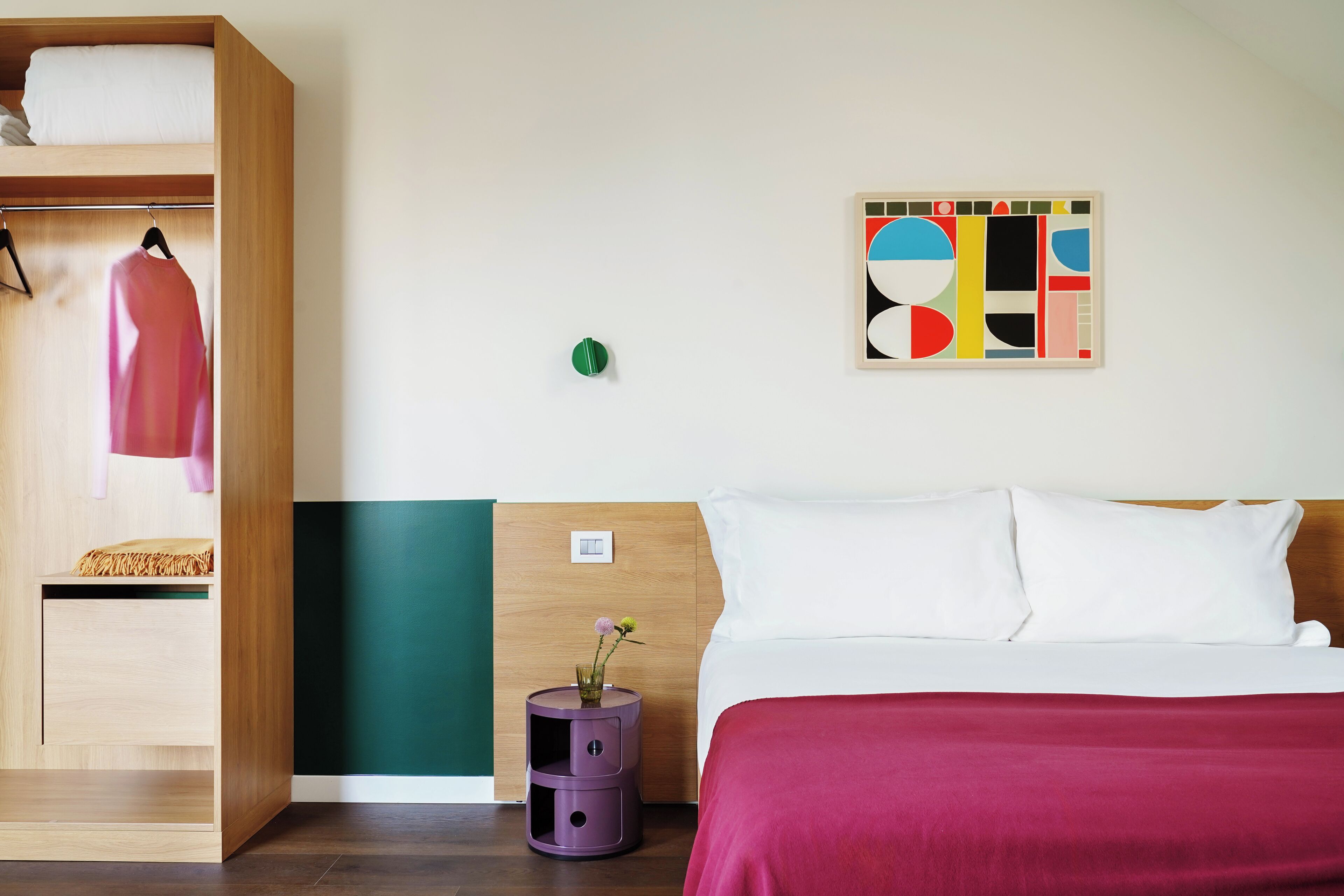 A hotel room with a bed, a wardrobe, a small table with a vase, and a colorful abstract painting on the wall.