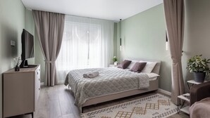 A bedroom with a bed, a TV on a cabinet, a bedside table with a plant, and a window with curtains.