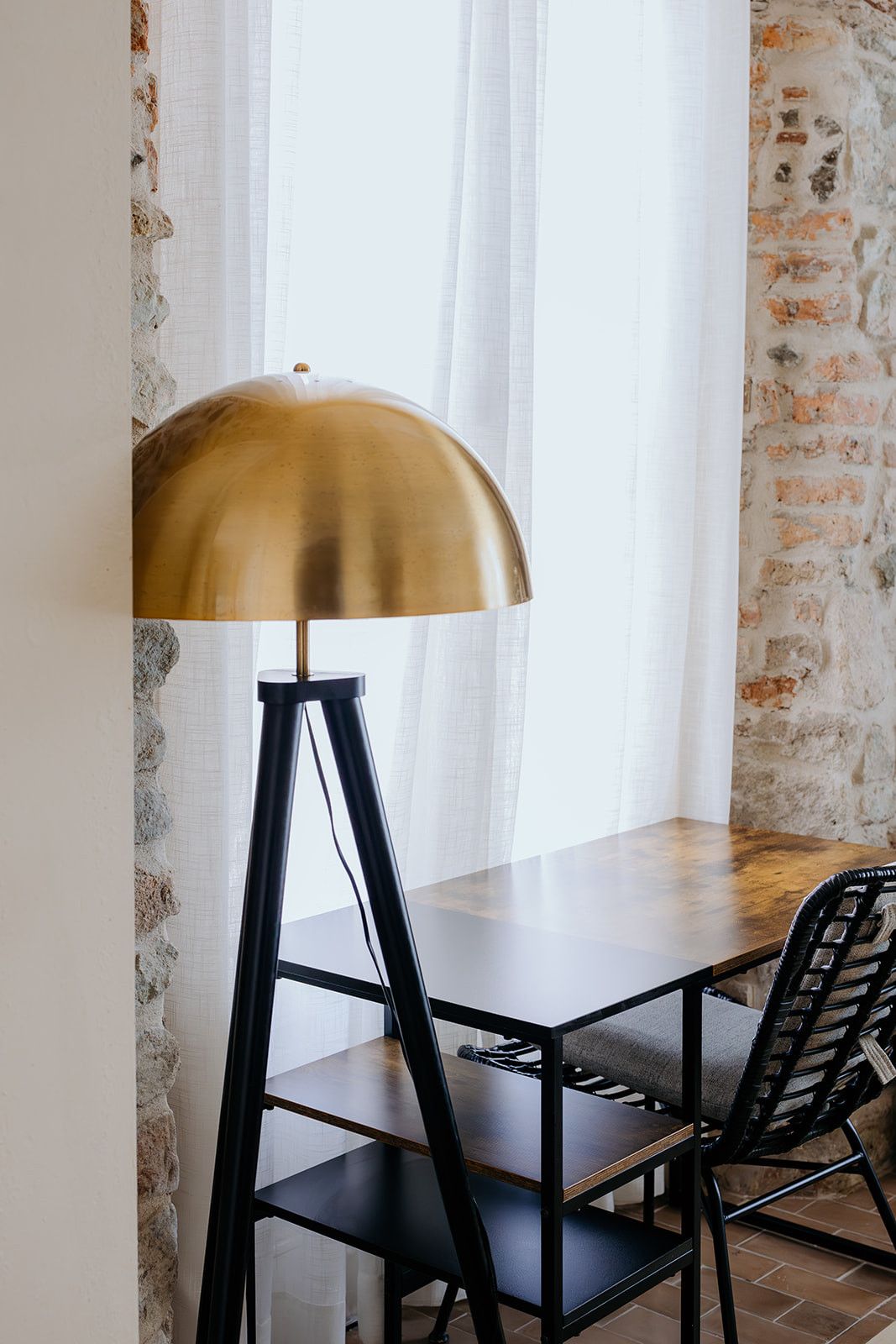 A table with a black metal frame and a single black chair. A gold-colored lamp with a dome shade is placed on the table. The background features a white curtain and a brick wall.