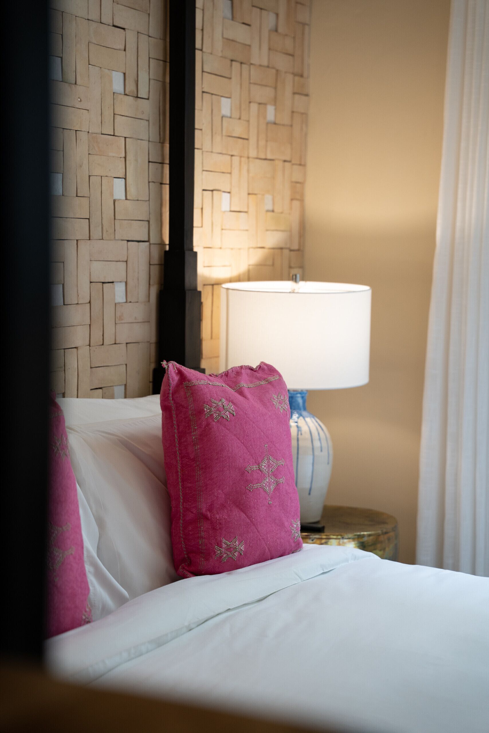 Un letto con lenzuola bianche e un cuscino rosa, una lampada su un comodino e un motivo decorativo intrecciato al muro.