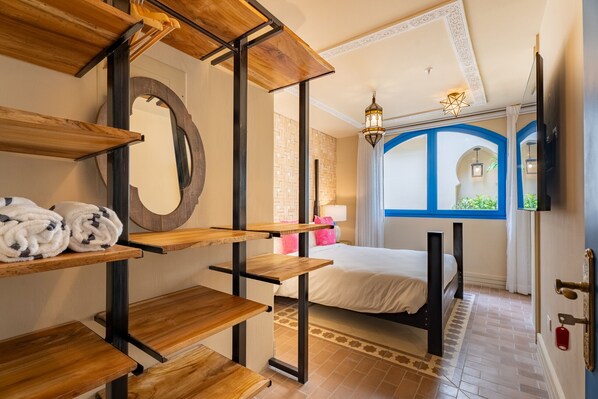 Une chambre d’hôtel avec un lit, des étagères en bois, un miroir et une vue sur l’extérieur par une fenêtre dotée d’un cadre bleu.