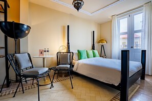 Ett hotellrum med en säng, två stolar, ett bord med toalettartiklar, en lampa och utsikt över byggnader genom fönstret.