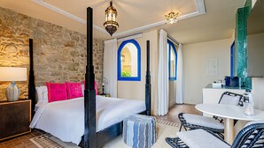 A bedroom with a stone wall, a bed with white bedding and pink pillows, a nightstand with a lamp, and a view through an arched window. - Balcony Superior with communal pool Ma7 (Ciudad de Panamá)