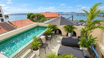A rooftop pool with a view of the sea, outdoor seating, and a bridge in the background.