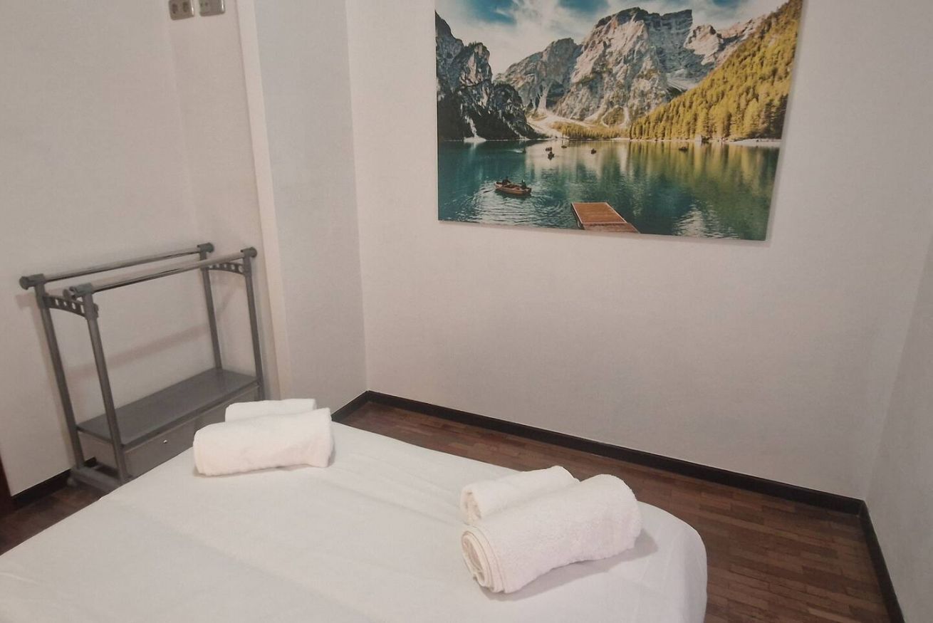 A hotel room with a bed, two rolled towels, a wooden floor, a metal rack, and a painting of a lake with mountains.