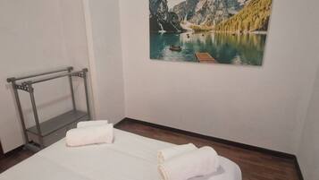 A hotel room with a bed, two rolled towels, a wooden floor, a metal rack, and a painting of a lake with mountains.