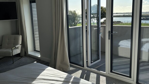 Ein Schlafzimmer mit einem großen Bett, einem Sessel und Blick auf ein Gewässer durch eine Glastür.