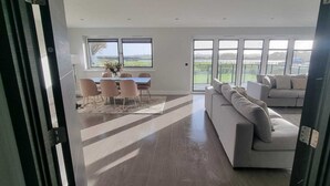 Ein großzügiger Wohnraum mit einer breiten Couch, einem Essbereich mit Stühlen und einem Tisch sowie großen Fenstern mit Blick ins Freie.