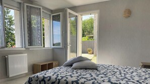 Un dormitorio con una cama grande, un radiador, un banco de madera y vistas a un jardín a través de ventanas abiertas.