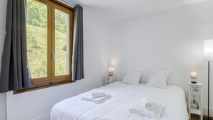 A bedroom with a large bed, two bedside tables with lamps, a window with curtains, and a view of greenery outside. - Rive Bleu: Charming T2 with Shared Beach (Doussard)