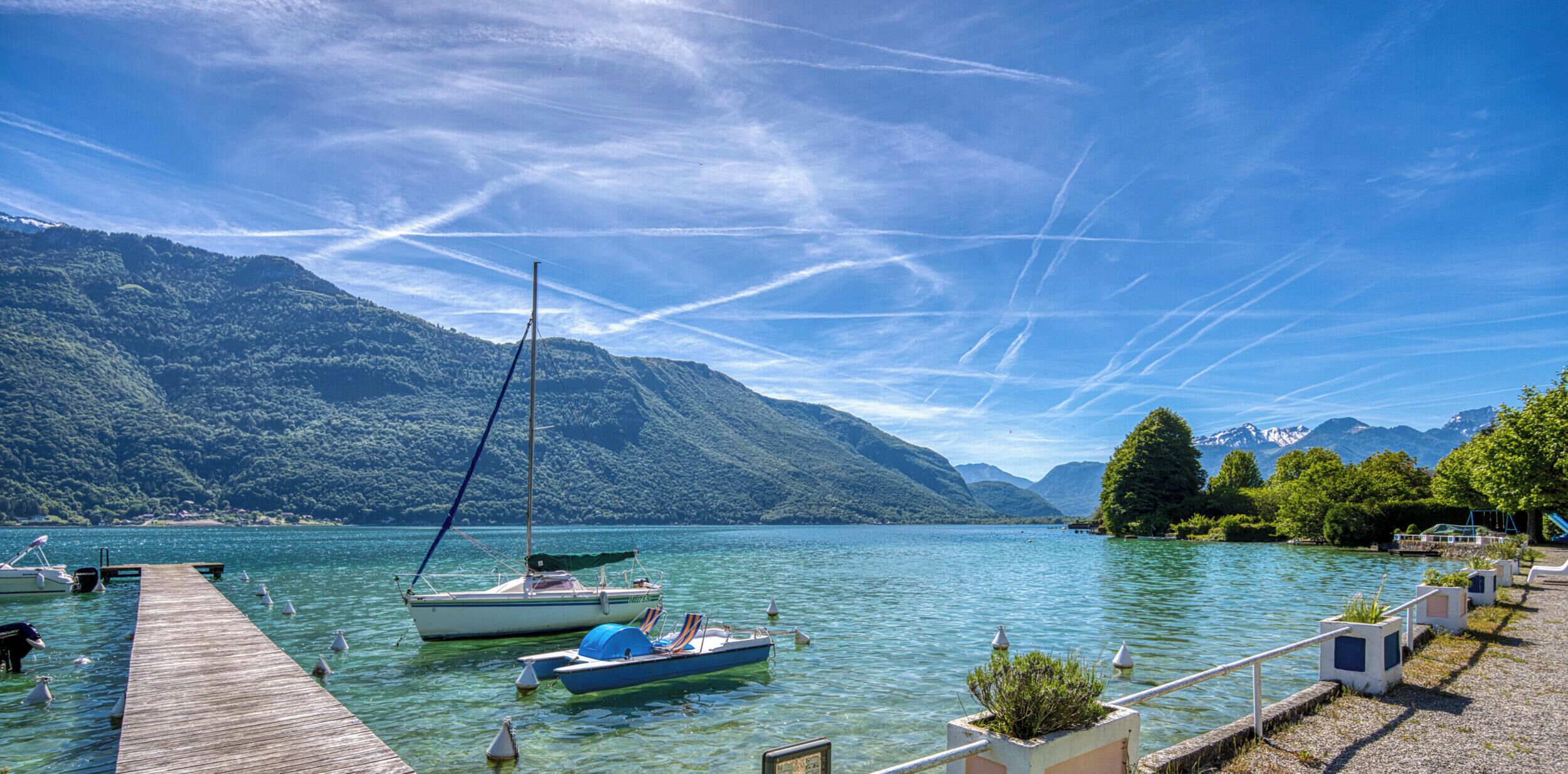 Ein ruhiger See mit Booten, einer hölzernen Anlegestelle und Bergen im Hintergrund.
