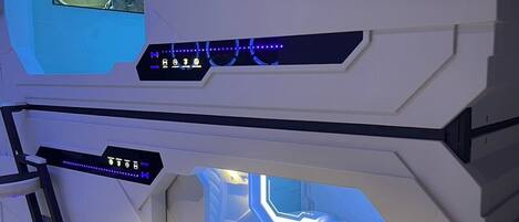 Une pièce futuriste équipée d’un panneau de contrôle, d’un lit et d’une fenêtre donnant sur un paysage urbain.