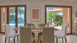 Un coin repas avec une table dressée pour quatre personnes, entourée de chaises, et offrant une vue sur une piscine et des palmiers à l’extérieur.