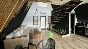 A modern loft-style living space with a sloped ceiling, a glass dining table, a sofa, and a kitchen area with a built-in stove. - Lost In Sky (Yenokavan)